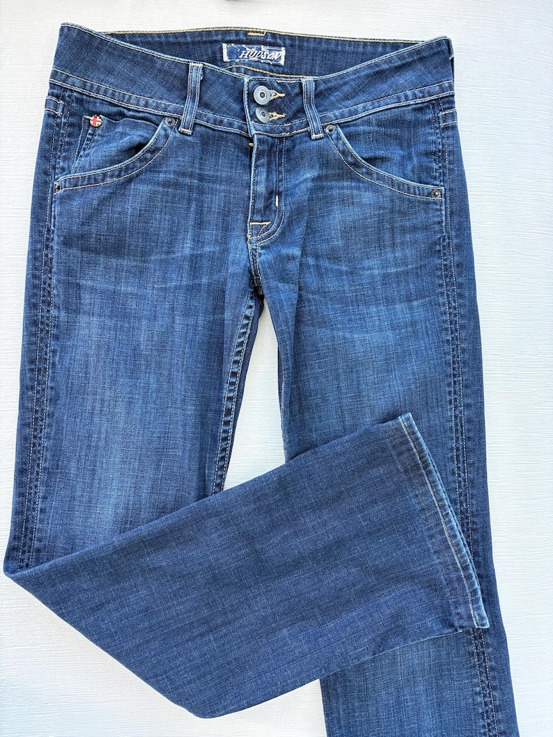 Może przedstawiać: Ciemnoniebieskie dżinsy z klasycznym pięciokieszeniowym designem. Dżinsy mają zapięcie na guzik i zamek błyskawiczny, szlufki na pasek i markę "Hudson" na pasku. Dżinsy są złożone, prezentując fakturę i pranie denimu.