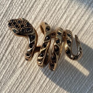 Op de afbeelding: Een goudkleurige ring in de vorm van een kronkelende slang. De kop van de slang is ingelegd met kleine, donkerzwarte steentjes en het lichaam heeft een reeks herhalende, getextureerde segmenten, elk ingelegd met een enkel zwart steentje.