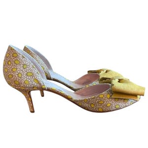Op de afbeelding: Een paar gele en witte pumps met madeliefjespatroon en een strikdetail. De schoenen hebben een spitse neus en een stilettohak. De hakken meten ongeveer 7.6 cm.