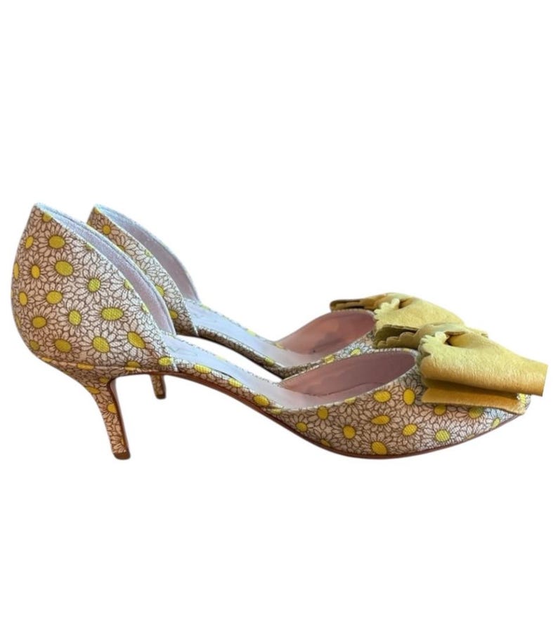 Op de afbeelding: Een paar gele en witte pumps met madeliefjespatroon en een strikdetail. De schoenen hebben een spitse neus en een stilettohak. De hakken meten ongeveer 7.6 cm.