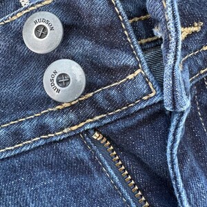 Może przedstawiać: Zbliżenie na ciemnoniebieskie dżinsy. Widoczne są dwa srebrne guziki z napisem "HUDSON", a także zamek błyskawiczny i kontrastowe szwy. Denim ma teksturowany wygląd.