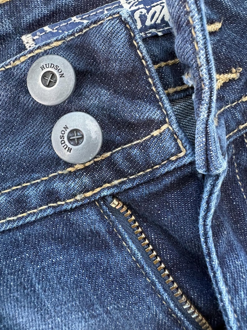 Może przedstawiać: Zbliżenie na ciemnoniebieskie dżinsy. Widoczne są dwa srebrne guziki z napisem "HUDSON", a także zamek błyskawiczny i kontrastowe szwy. Denim ma teksturowany wygląd.