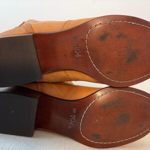 May include: A pair of light brown leather boots, viewed from the soles up. The soles are a darker brown with white stitching around the edges. The heels are black. The text "No 704" is visible on the sole.