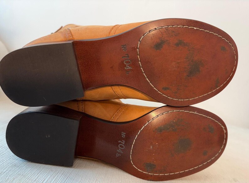 May include: A pair of light brown leather boots, viewed from the soles up. The soles are a darker brown with white stitching around the edges. The heels are black. The text "No 704" is visible on the sole.