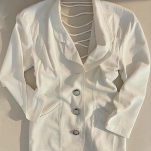Puede incluir: Un blazer blanco con un dise&ntilde;o &uacute;nico de espalda abierta. El blazer presenta un cuello con muescas, mangas largas y tres botones plateados en la parte delantera. La espalda tiene una serie de correas verticales. La chaqueta est&aacute; hecha de una tela lisa, posiblemente tejida.