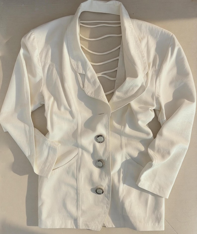 Puede incluir: Un blazer blanco con un dise&ntilde;o &uacute;nico de espalda abierta. El blazer presenta un cuello con muescas, mangas largas y tres botones plateados en la parte delantera. La espalda tiene una serie de correas verticales. La chaqueta est&aacute; hecha de una tela lisa, posiblemente tejida.