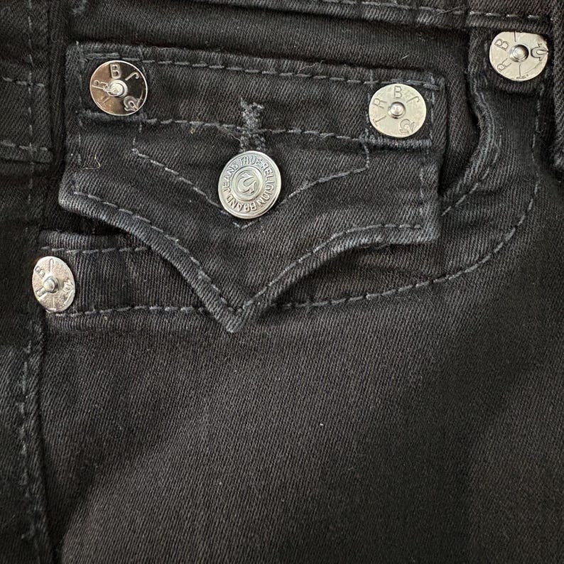 Puede incluir: Primer plano de unos vaqueros negros de mezclilla con botones de cierre de metal plateado y detalles de bolsillo. Los botones est&aacute;n grabados con un logotipo. Las costuras resaltan el dise&ntilde;o del bolsillo.