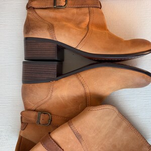 May include: Two light brown leather ankle boots with a buckle closure and a stacked heel. The boots have a rounded toe and a dark brown sole. The heels are approximately 2 inches high.