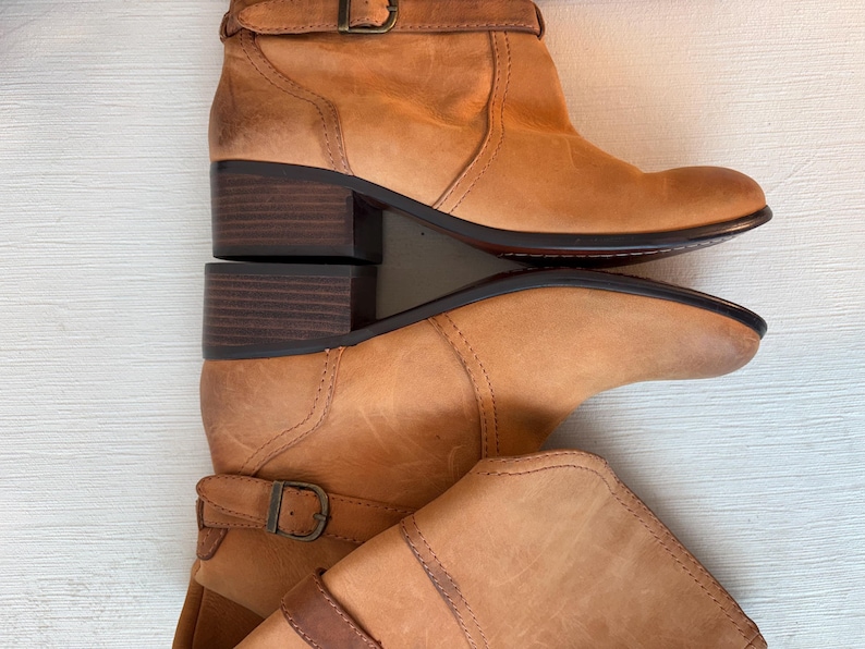 May include: Two light brown leather ankle boots with a buckle closure and a stacked heel. The boots have a rounded toe and a dark brown sole. The heels are approximately 2 inches high.