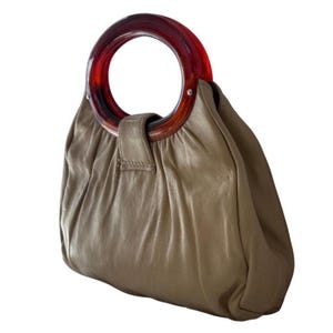 Puede incluir: Un bolso de mano color topo con un asa circular de color marrón rojizo. El bolso tiene un diseño fruncido y un pequeño cierre de correa. El asa es de un material translúcido y el bolso parece ser de cuero.