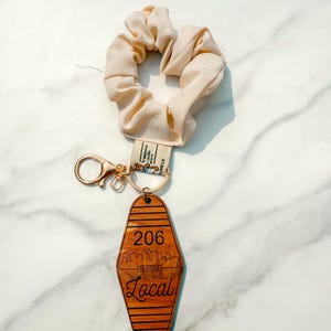 May include: A beige scrunchie and a wooden keychain with a gold-colored clasp. The keychain is shaped like a motel key, with the number "206" and the word "Local" below a Seattle skyline design.