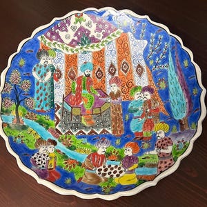 Puede incluir: Un plato de cerámica decorativo con borde festoneado, que presenta una escena colorida pintada a mano. La obra de arte representa figuras con vestimenta tradicional, sobre un fondo de tienda de campaña y paisaje, con borde azul y ribete blanco.