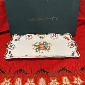May include: A rectangular serving tray with a floral and fruit design, gold trim, and gold handles. The tray is white with a scalloped edge. The box behind the tray has the text "I. Godinger & Co."