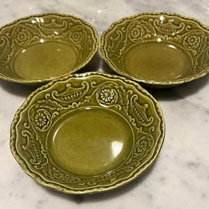 May include: Three olive green ceramic bowls with ornate, embossed floral and scrollwork designs around the rims. The bowls have a scalloped edge and a shallow, rounded interior. The bowls are arranged on a light-colored surface.