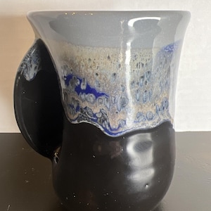 May include: A handmade ceramic mug with a unique design. The mug features a black base, transitioning to a blue, beige, and gray speckled middle, and a gray rim. The handle is black and has a distinctive shape.