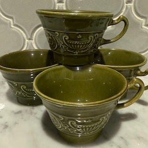 May include: Four olive green ceramic teacups with handles and decorative embossed floral patterns. The cups are stacked and arranged on a white marble surface, showcasing their design and color.