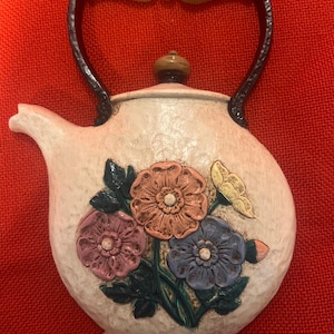 May include: Cream-colored ceramic teapot with a floral design. The teapot features a brown handle and spout, with a lid and a decorative bouquet of flowers in shades of pink, orange, blue, and yellow. The teapot is likely a decorative item.