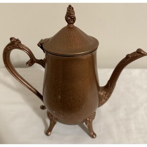 May include: A vintage copper-colored teapot with ornate detailing. The teapot features a curved handle, a long spout, and a decorative finial on the lid. It stands on three small, decorative feet, showcasing an antique aesthetic.