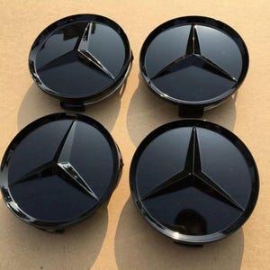 May include: Four black car wheel center caps with a three-pointed star emblem. The caps are circular with a glossy finish, and the star is raised and matte. The caps are arranged in a square formation.
