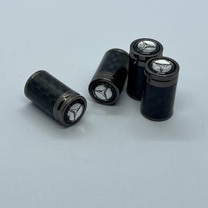 Op de afbeelding: Vier zwarte ventieldopjes met een koolstofvezelpatroon en een zilveren embleem. De doppen hebben een cilindrische vorm met een donkere metalen basis en bovenkant. Het embleem heeft een driepuntige ster.