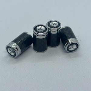 Op de afbeelding: Vier zwarte en zilveren ventieldopjes voor banden. De doppen hebben een koolstofvezelpatroon op de cilindrische behuizing en een zilveren bovenkant met een Mercedes-Benz logo. De doppen zijn op een wit oppervlak geplaatst.