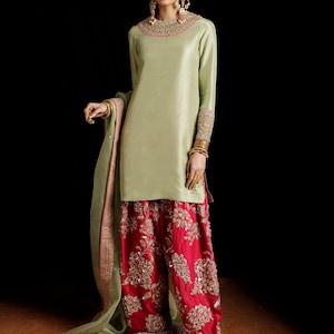 May include: A stylish outfit comprising a pale green tunic with long sleeves and a round neck, complemented by striking red palazzo trousers embellished with gold embroidery. The ensemble includes a matching green scarf and gold jewellery.