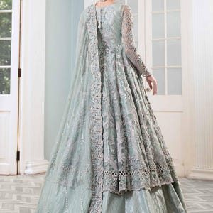 May include: A pale green formal gown with detailed embroidery and a flowing cape. The dress has long sleeves and a layered skirt, creating an elegant silhouette. The gown is embellished with delicate silver accents.