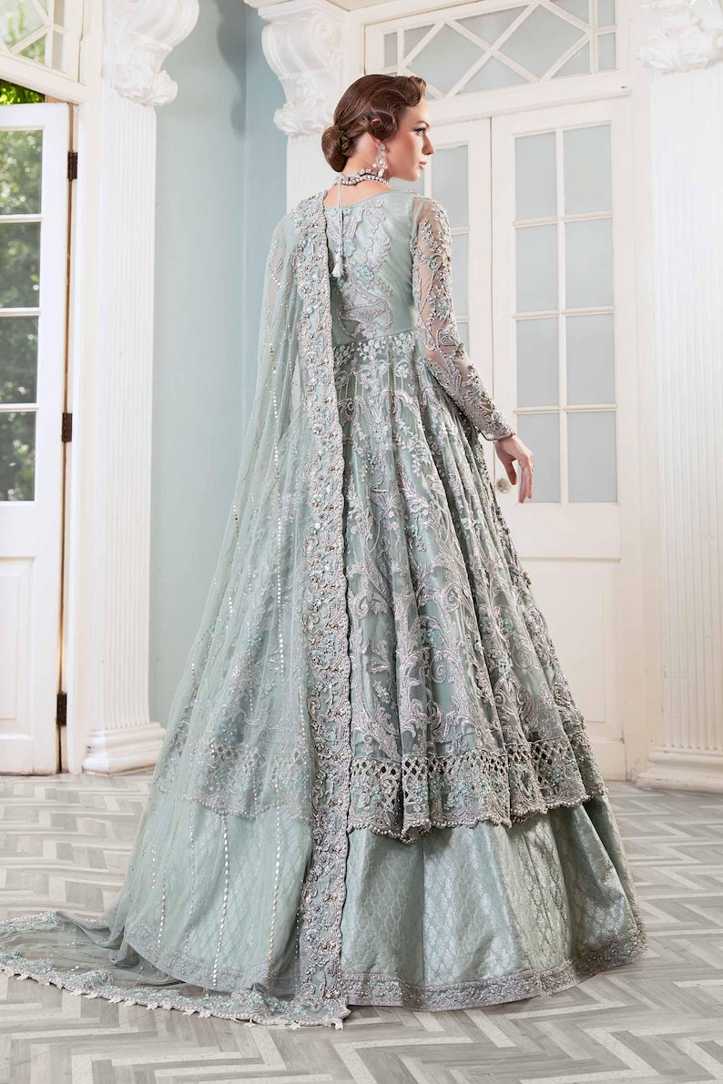 May include: A pale green formal gown with detailed embroidery and a flowing cape. The dress has long sleeves and a layered skirt, creating an elegant silhouette. The gown is embellished with delicate silver accents.