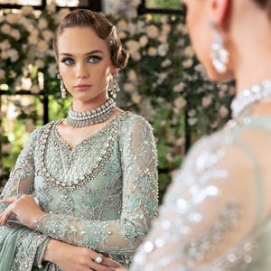 May include: A woman in a pale green, elaborately embroidered gown with long sleeves, a beaded neckline, and matching jewellery. The dress features floral embroidery and is reflected in a mirror. The background is blurred with white flowers.