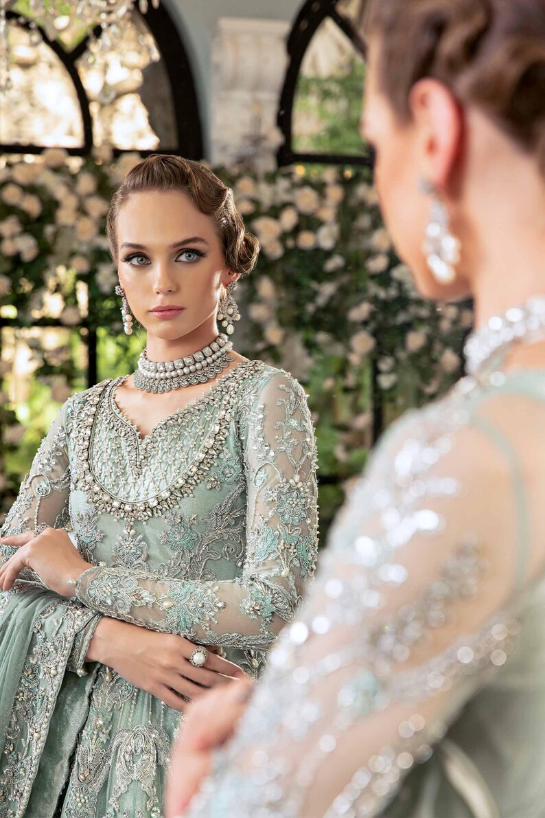 May include: A woman in a pale green, elaborately embroidered gown with long sleeves, a beaded neckline, and matching jewellery. The dress features floral embroidery and is reflected in a mirror. The background is blurred with white flowers.
