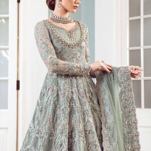 May include: A long, sage green formal gown with intricate silver embroidery and beading. The dress features long sleeves, a V-neckline, and a full skirt. A matching shawl is draped over the arm. The gown is suitable for special occasions.