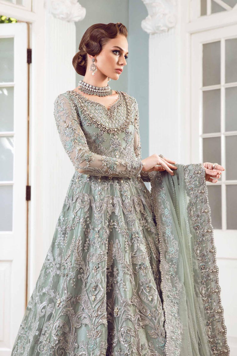May include: A long, sage green formal gown with intricate silver embroidery and beading. The dress features long sleeves, a V-neckline, and a full skirt. A matching shawl is draped over the arm. The gown is suitable for special occasions.