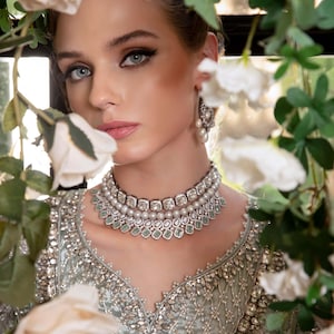 May include: A woman wearing a silver and pearl necklace and matching earrings. She is also wearing a light green, intricately embroidered top. White roses and green foliage frame the subject.