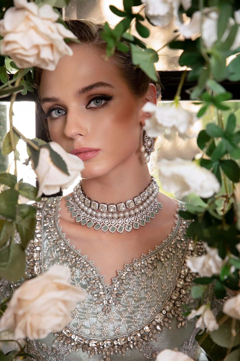 May include: A woman wearing a silver and pearl necklace and matching earrings. She is also wearing a light green, intricately embroidered top. White roses and green foliage frame the subject.