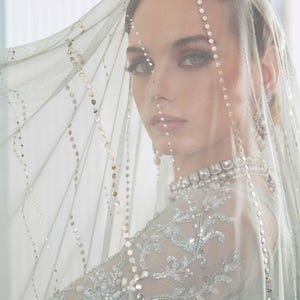 May include: A woman in a light blue wedding dress with detailed silver embroidery and a sheer veil decorated with sequins. The veil drapes over the face, creating a soft effect. The dress has a high neckline and long sleeves.