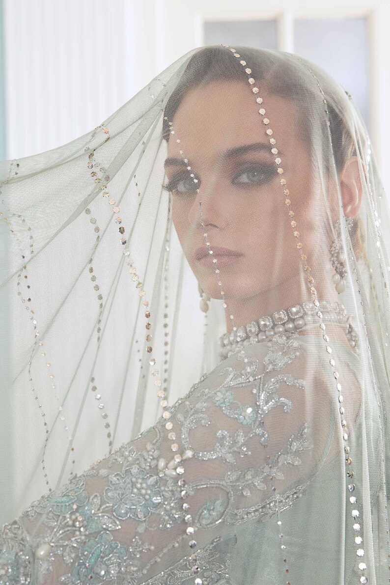 May include: A woman in a light blue wedding dress with detailed silver embroidery and a sheer veil decorated with sequins. The veil drapes over the face, creating a soft effect. The dress has a high neckline and long sleeves.