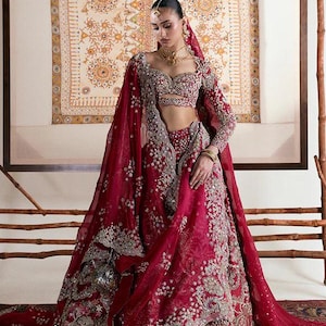 May include: A woman in a red bridal ensemble. The outfit includes a long-sleeved top, a full skirt, and a sheer head covering. The outfit is heavily embellished with silver embroidery and beadwork. The background features a framed tapestry.