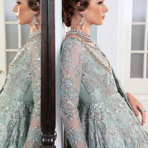 May include: A pale teal formal gown with detailed silver embroidery and beading. The long-sleeved dress has a floral pattern and a flowing skirt. The model is wearing a matching necklace and earrings, with her hair styled in an updo.