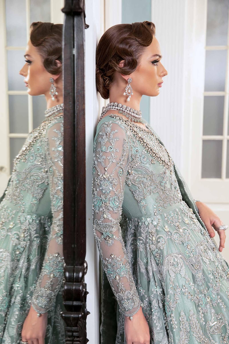 May include: A pale teal formal gown with detailed silver embroidery and beading. The long-sleeved dress has a floral pattern and a flowing skirt. The model is wearing a matching necklace and earrings, with her hair styled in an updo.