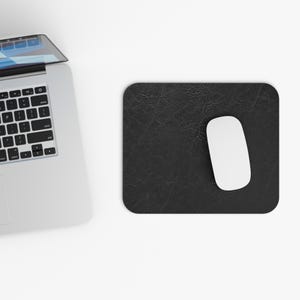 May include: A black rectangular mouse mat with a textured finish and rounded corners. A white wireless mouse rests on the mat. A laptop is partially visible in the top left corner.