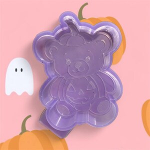 以下が含まれることがあります： ハロウィーンをテーマにした紫色のプラスチック製のクッキーカッター。カボチャの顔をしたクマの形です。白い幽霊とオレンジ色のカボチャが背景にあります。お祝いのお菓子作りに最適です。