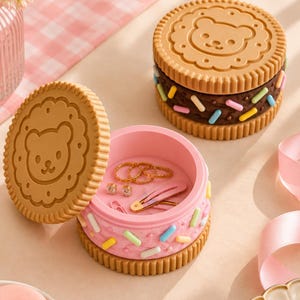 Puede incluir: Dos recipientes de almacenamiento redondos con forma de galleta y caras de oso en relieve en las tapas. Un recipiente está abierto, mostrando anillos dorados, pendientes y una pinza para el pelo rosa. Ambos tienen lados texturizados con chispas.
