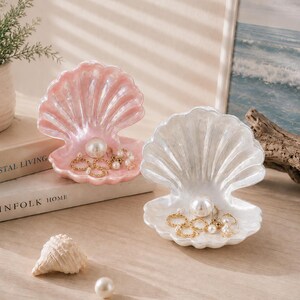Può includere: Due piatti portagioie a forma di conchiglia aperta, uno rosa iridescente e uno bianco iridescente, contenenti ciascuno una grande perla artificiale e diversi anelli dorati. Una piccola conchiglia e una perla artificiale sono poste davanti.