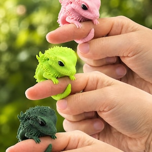 Może przedstawiać: Trzy małe, teksturowane figurki jaszczurek w kolorze różowym, limonkowym i ciemnozielonym. Każdy miniaturowy gad ma kolczasty grzbiet, długi ogon owijający się wokół palca oraz duże, gładkie czarne oczy, wykończone matową powierzchnią.