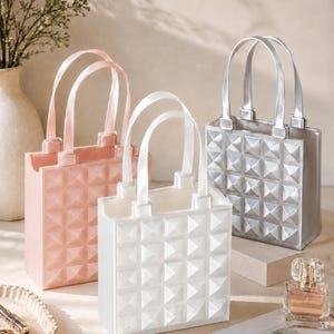 May include: Three rectangular tote bags in light pink, white, and silver. Each bag features a raised pyramid stud pattern on the exterior, two vertical handles, and an open top. The bags are arranged in a cluster, showcasing their structured design.