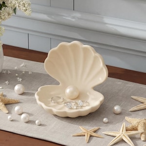 Pode incluir: Uma concha de cerâmica creme contém anéis e uma grande pérola. Estrelas do mar e pérolas de vários tamanhos estão espalhadas sobre um corredor neutro. Um vaso branco com flores está ao fundo.