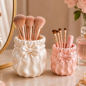 Peut inclure: Deux porte-pinceaux de maquillage cylindriques avec une texture plissée rappelant le tissu et un nœud central. Le plus grand pot est de couleur crème et contient trois gros pinceaux, tandis que le plus petit est rose pâle et contient quatre pinceaux.