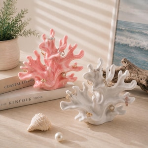 May include: Two coral-shaped jewellery holders, one in glossy pastel pink and one in white, featuring multiple branches for hanging rings. The holders are displayed alongside a small white seashell and a single white pearl on a flat surface.