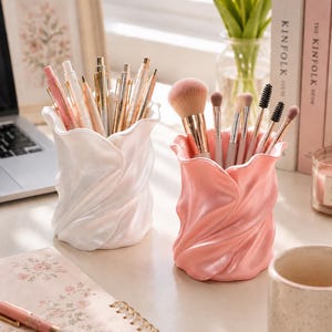May include: Two cylindrical desk organisers with a ruffled, petal-like texture. One organiser is pearlescent white and holds various pens, while the other is a soft, glossy pink and contains a collection of makeup brushes with metallic handles.
