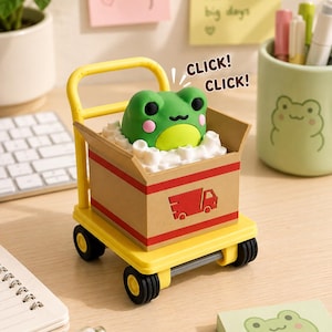 May include: A yellow plastic cart with black wheels carrying a brown cardboard box. Inside the box, a green frog figure with pink cheeks sits on white packing material. The box features a red truck icon and the text CLICK! CLICK! in black.